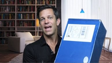 Man speaking excitedly while holding a large blue package with a shipping label in a living room
