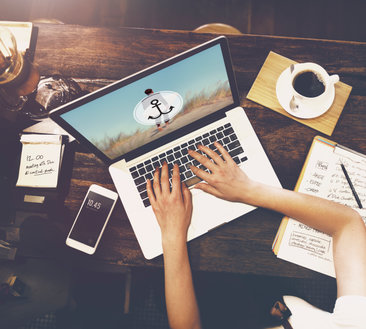Hands typing on a laptop displaying a beach graphic, with coffee and notebooks on a wooden desk