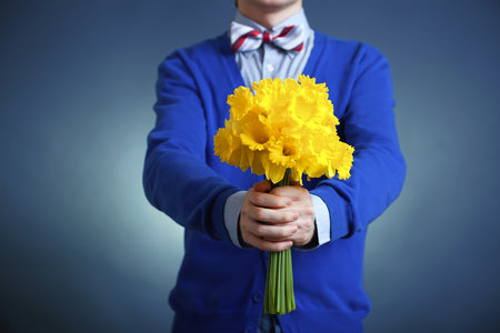 Man in a blue cardigan and striped bow tie holds out a bouquet of yellow daffodils