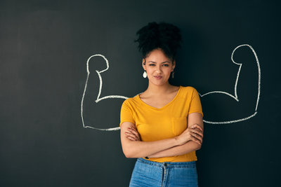Woman with arms crossed stands confidently in front of chalk-drawn muscular arms on a blackboard