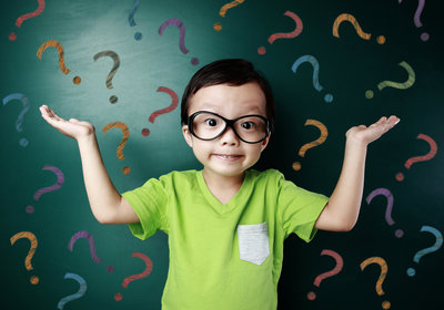 Confused young boy in glasses shrugs with hands raised against a chalkboard covered in question marks