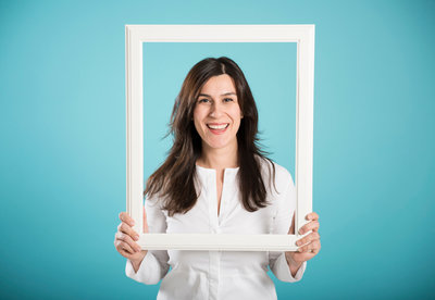 Smiling woman in a white shirt holds an empty white picture frame around her face against a teal background