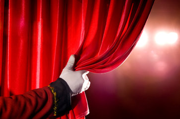 A gloved hand in formal attire pulls open a red velvet curtain, revealing bright stage lights behind it.