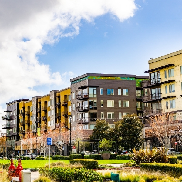 Multistory apartment building exterior with balconies, landscaped courtyard, and green lawn under a blue sky