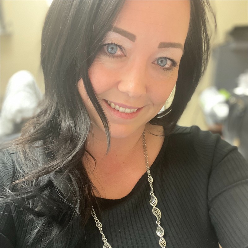 Woman with dark wavy hair and a silver necklace smiles for a close-up selfie indoors