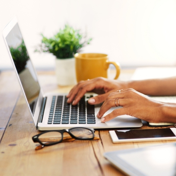 Hands typing on a laptop keyboard beside glasses, a mug, and a small plant on a wooden desk