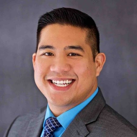 Smiling man in a gray suit and blue shirt with a patterned tie against a dark background